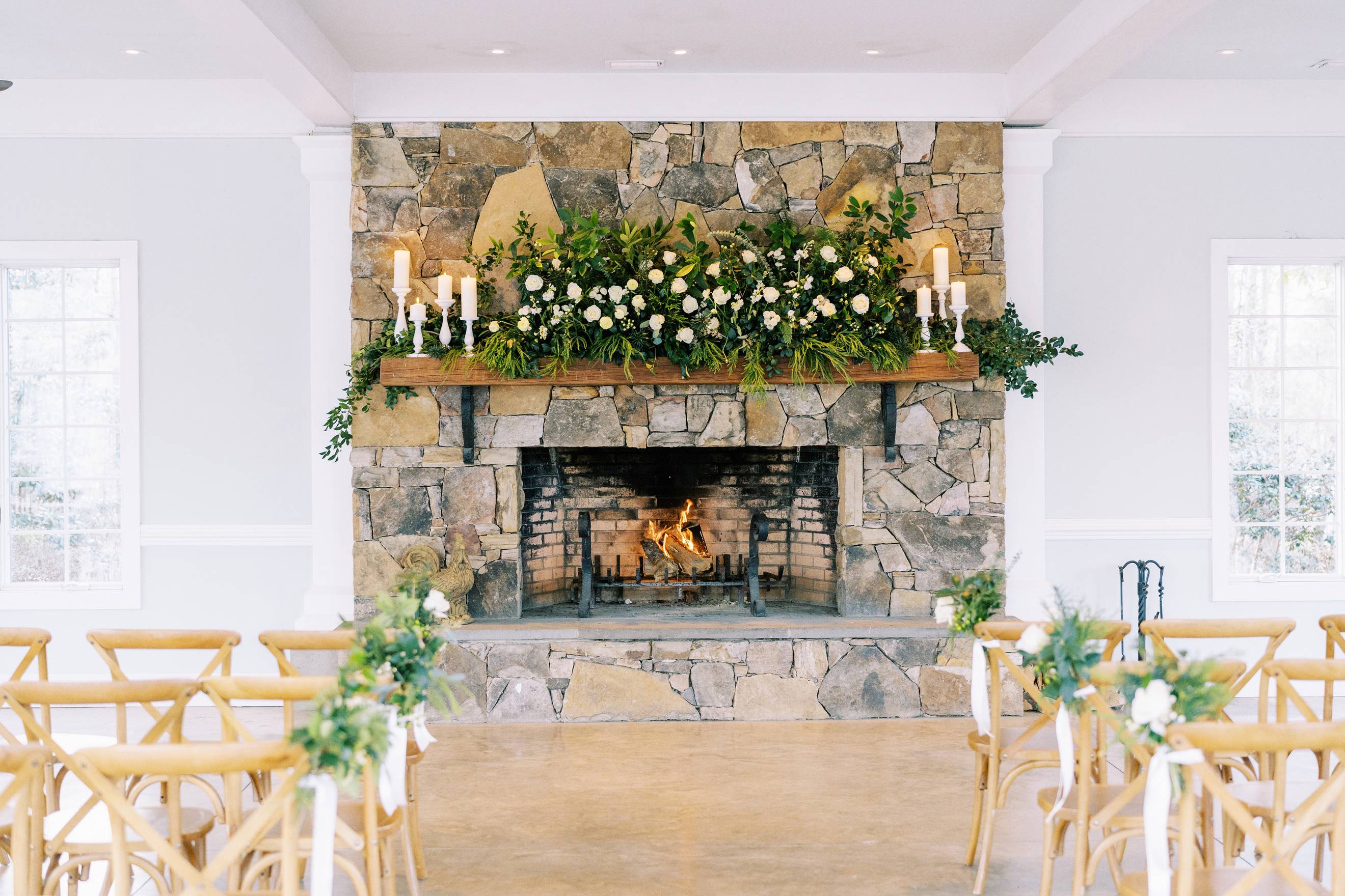 A stone fireplace decorated with green foliage, white flowers, and white candles, with a fire burning inside. Wooden chairs with floral arrangements are arranged in rows facing the fireplace.