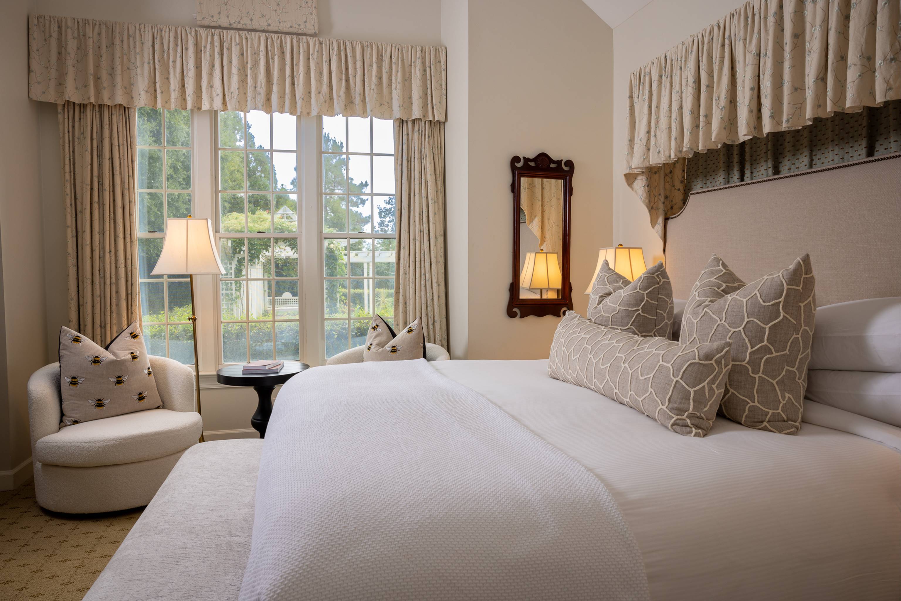 A bright, elegant bedroom with a large bed featuring patterned pillows, a cozy armchair with cushions, two tall lamps, a side table, a wall mirror, and large windows with drapes letting in natural light.