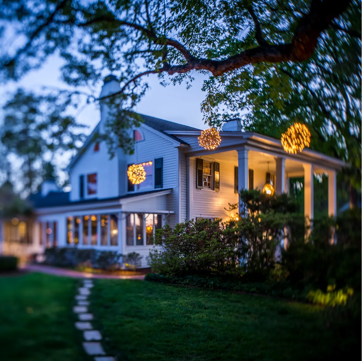 A white house with a wraparound porch is decorated with glowing orb lights hanging from trees. The yard is green with a stone path leading to the entrance, and the scene is set at dusk.