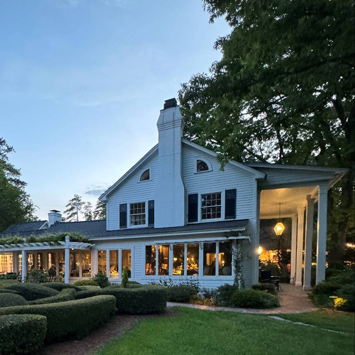 A large white house with dark shutters and a tall chimney, surrounded by neatly trimmed hedges and trees, is softly illuminated at dusk. Warm lights glow from the windows and a covered porch with a hanging lantern.