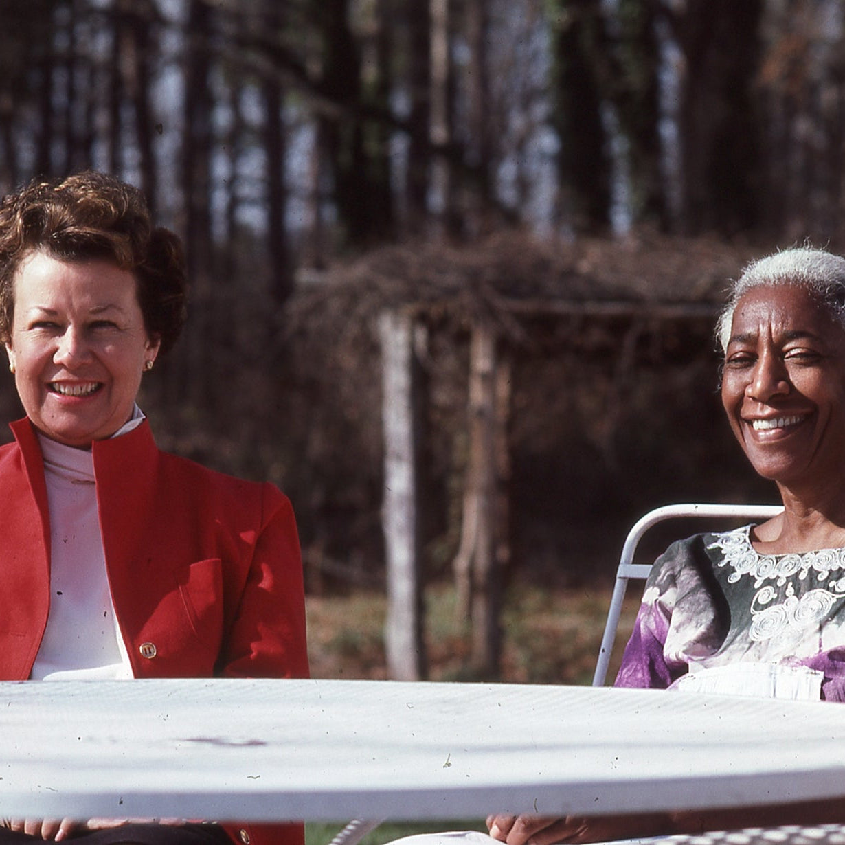 Two older women sit outdoors at a white table, smiling. One wears a red jacket over a pink blouse, the other wears a purple and white dress. Leafless trees and a rustic wooden arbor are visible in the background.
