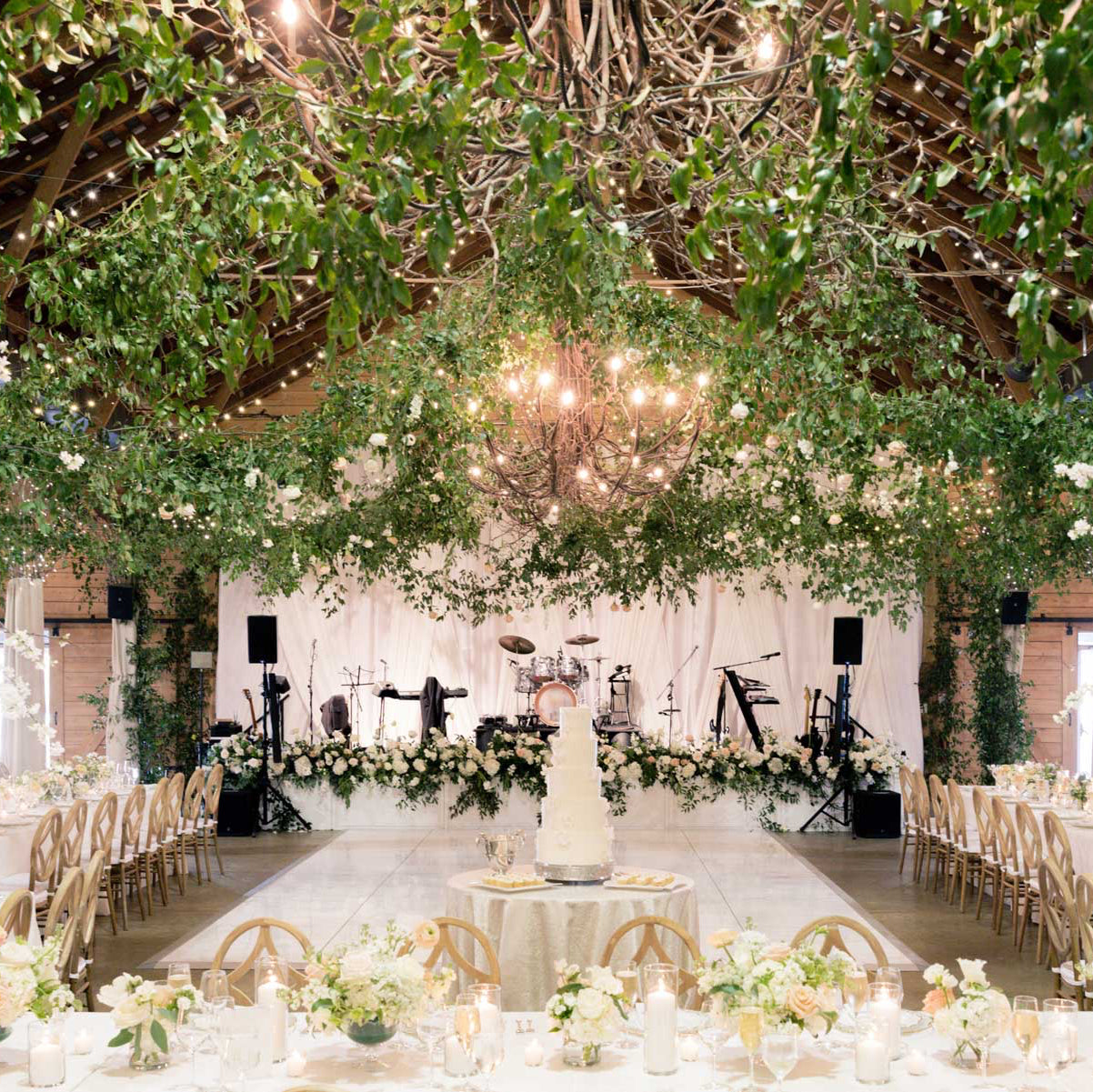 A beautifully decorated wedding reception venue with hanging greenery, chandeliers, white floral arrangements, a stage with musical instruments, and an elegant white wedding cake in the center.