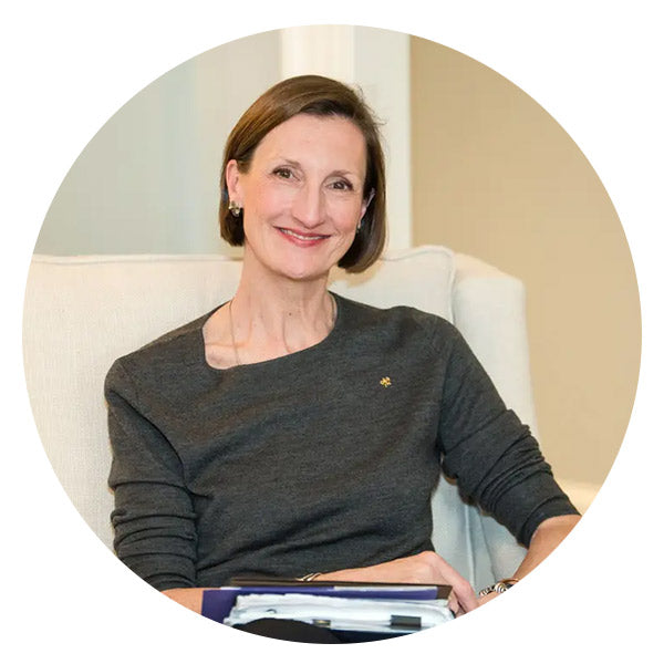 A woman with short brown hair and a dark grey top sits on a light-colored couch, smiling at the camera. She is holding a stack of folders or papers on her lap.
