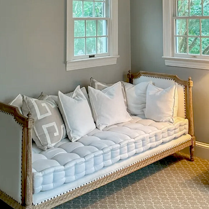 A wooden-framed daybed with a tufted white cushion and five white pillows sits against a light gray wall between two windows in a carpeted room. One pillow has a beige geometric pattern.