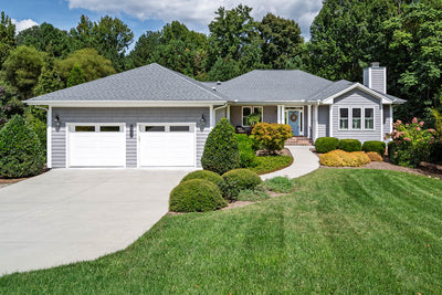 A single-story gray house with a well-kept front lawn, landscaped bushes, two white garage doors, and a curved driveway leading to a covered front porch. Trees and greenery surround the property.