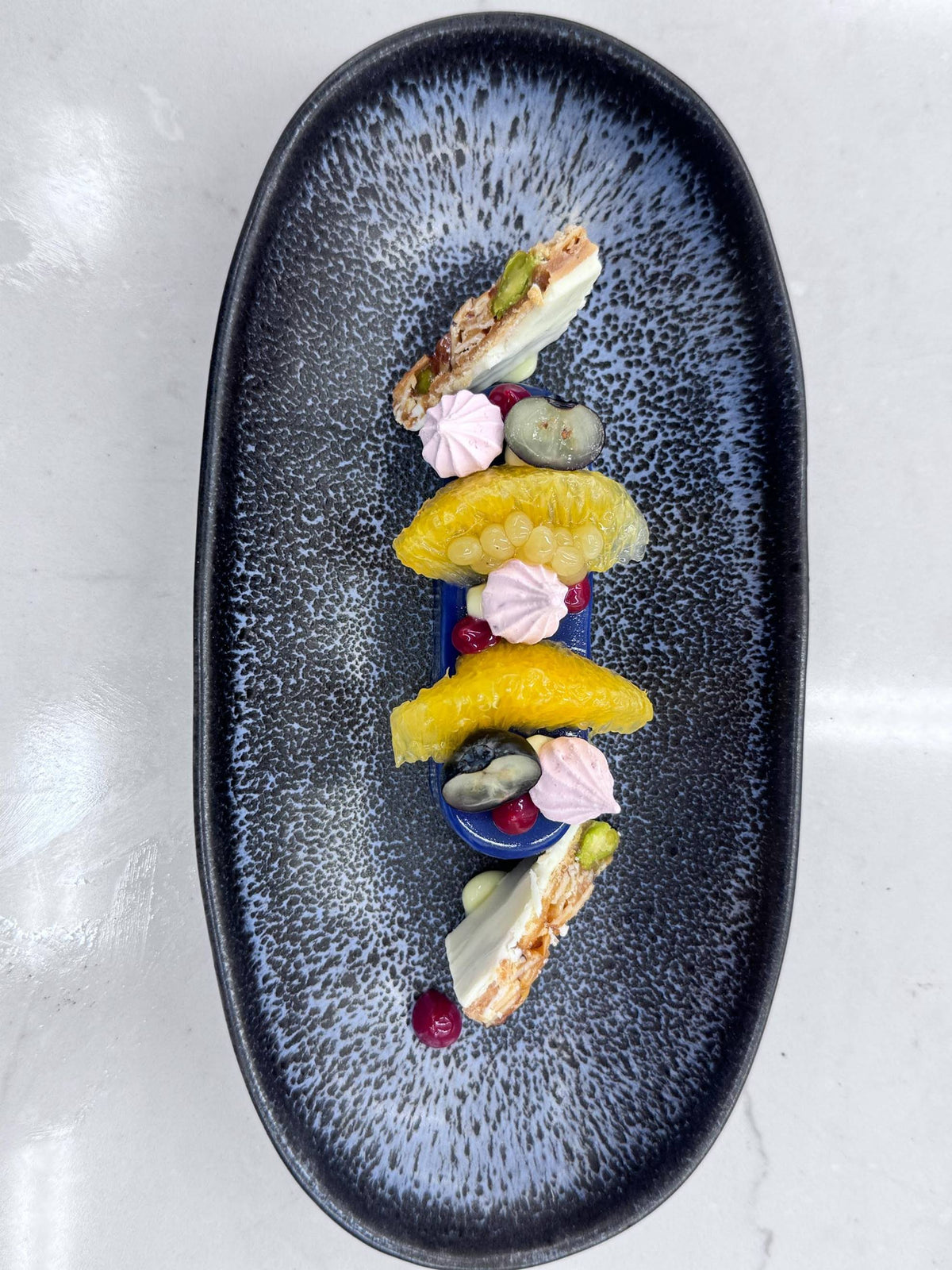 A black oval plate with a modern dessert featuring orange segments, pink meringue kisses, purple sauce, white chocolate shards, pistachios, and red berries, arranged artfully on a speckled blue background.