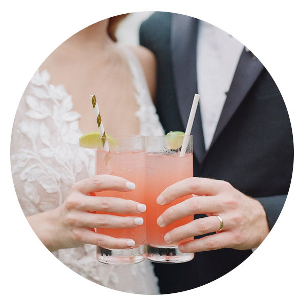A couple in formal attire holds two pink cocktails with lime wedges and striped straws, standing close together. The woman's dress is lacy and white, and the man's suit is dark with a white shirt.