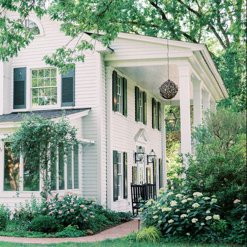 A white two-story house with black shutters and a covered front porch, surrounded by lush green plants and flowering bushes. A black porch swing hangs near the entrance, and large trees provide shade overhead.