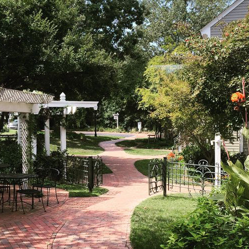 A winding brick pathway curves through a garden with green trees, flowering plants, an iron gate, and a white pergola with a black metal table and chairs on the left. A house is partially visible on the right.