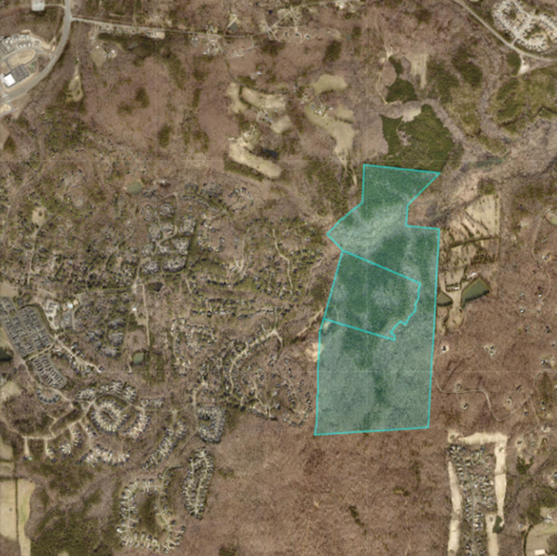 Aerial view of a wooded, suburban area with residential neighborhoods surrounding a large, irregularly shaped plot of land outlined in blue near the center and right side of the image.