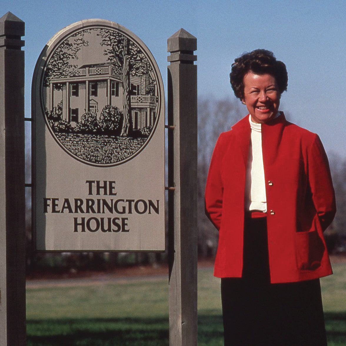A woman in a red jacket and white blouse stands smiling next to a large sign that reads "The Fearrington House" with a drawing of a house above the text. The background is outdoors with grass and a clear blue sky.