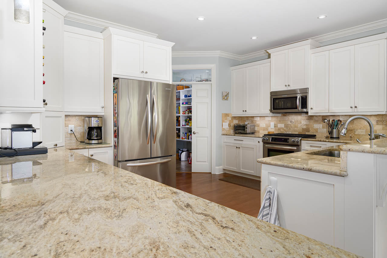 Modern kitchen with white cabinets, stainless steel appliances, beige granite countertops, a coffee maker, and a view of a pantry with shelves of food through an open door. Warm lighting creates a clean and inviting atmosphere.
