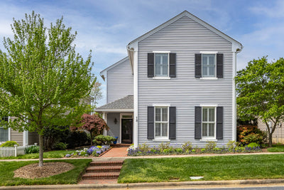 A two-story gray house with white trim and dark shutters, a brick walkway leading to the front door, neatly trimmed lawn, blooming flower beds, and trees on either side on a sunny day.