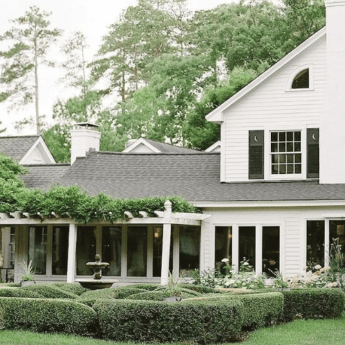 A white two-story house with black shutters, large windows, and a pergola over a patio. In front, manicured hedges form a geometric garden with a small fountain, surrounded by lush greenery and tall trees.