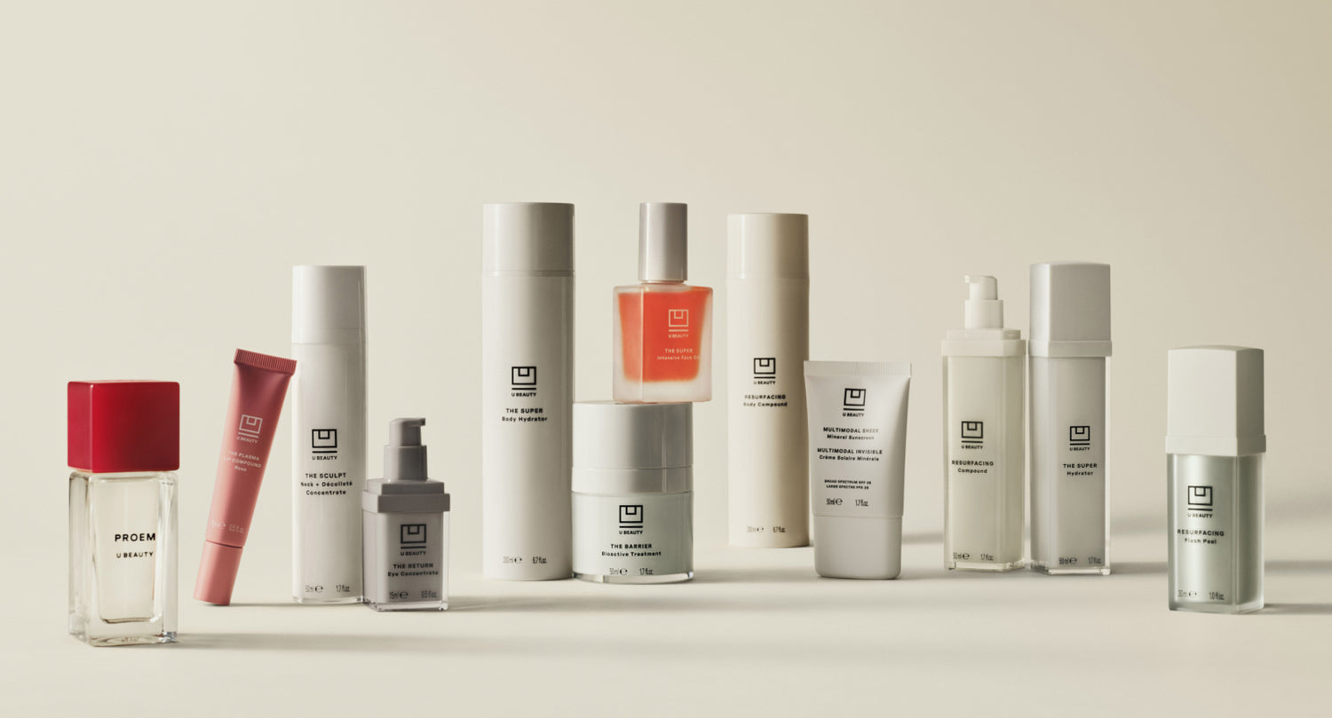 A collection of skincare products in minimalist white and clear bottles, tubes, and jars with black text. One perfume bottle has a red cap and label. The items are arranged on a neutral background.