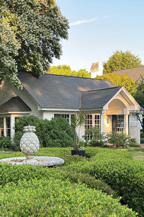 A white house with a gray roof and large windows is partially hidden by green bushes and trees. In the foreground, there is a decorative pineapple-shaped stone fountain surrounded by neatly trimmed hedges.