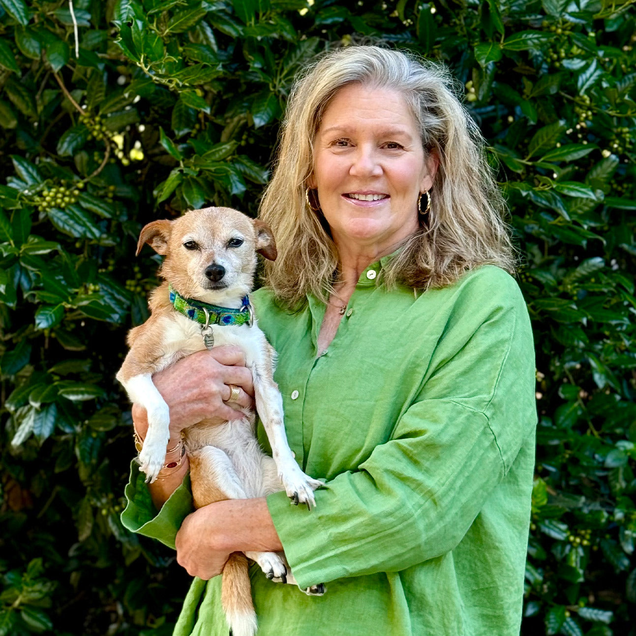A woman in a bright green shirt stands outdoors in front of leafy greenery, smiling and holding a small brown and white dog in her arms.