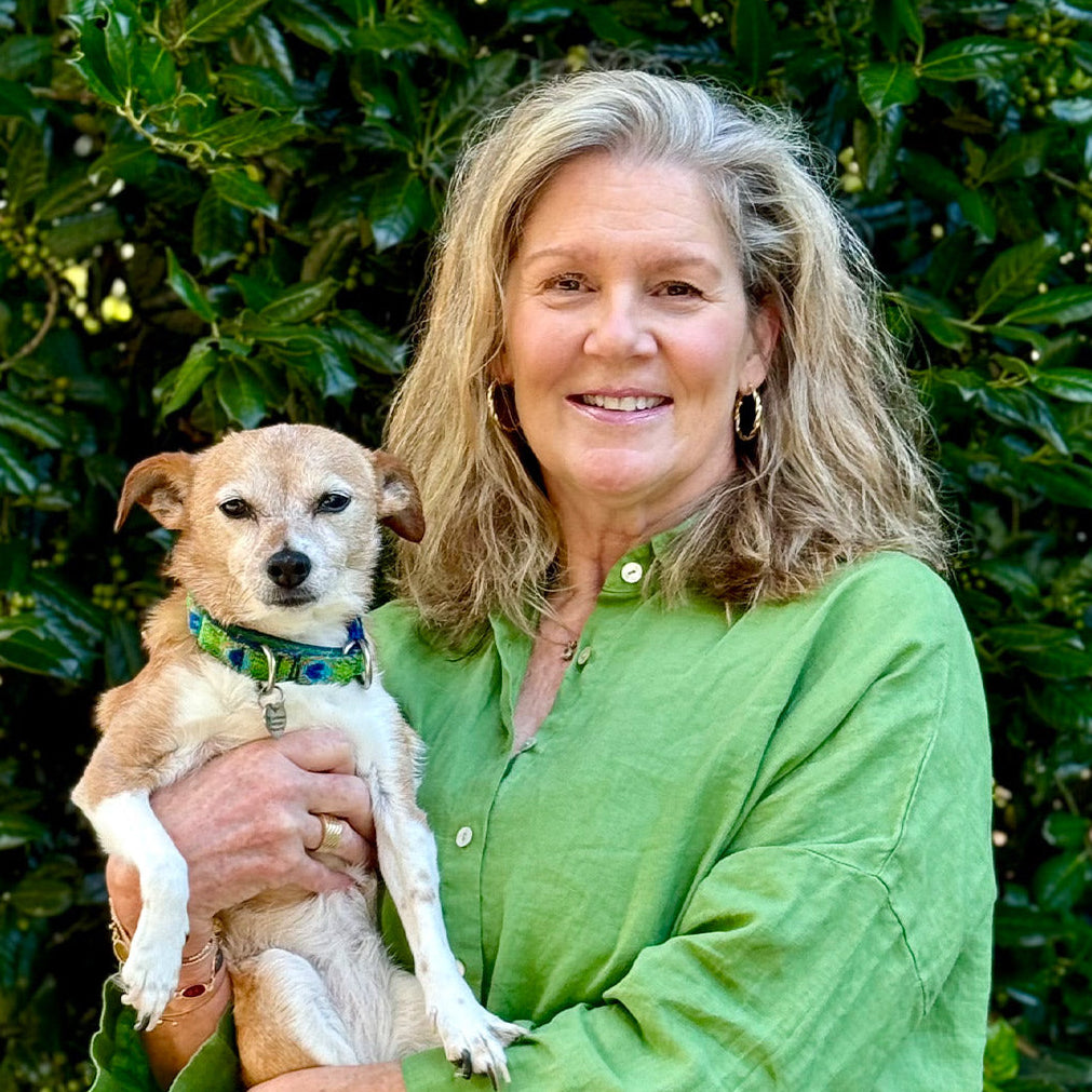 A smiling woman with wavy blond hair wearing a green shirt holds a small brown and white dog. They stand in front of lush green foliage.