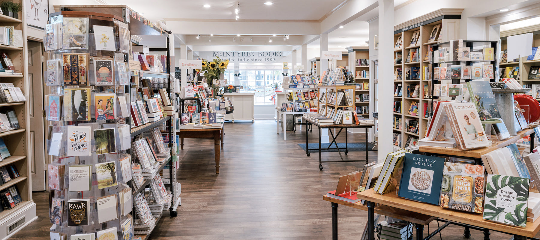 A bright, spacious bookstore with wooden floors, shelves and tables filled with books, and greeting cards on display racks. Signs and sunflowers decorate the entrance at the back, where natural light streams in.
