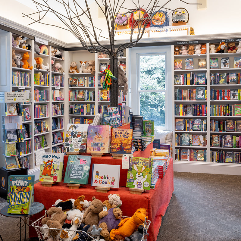 A brightly lit children’s bookstore features shelves full of colorful books and plush toys, a display table with picture books, and a decorative tree centerpiece. Large windows let in natural light.