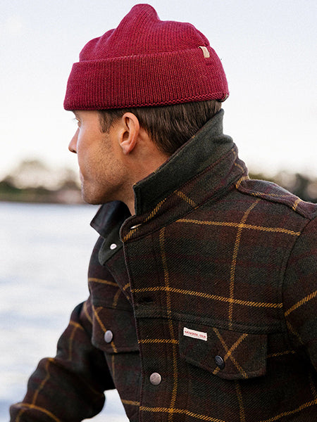 A man wearing a red knit beanie and a dark plaid wool jacket looks to the side, standing outdoors near a body of water.