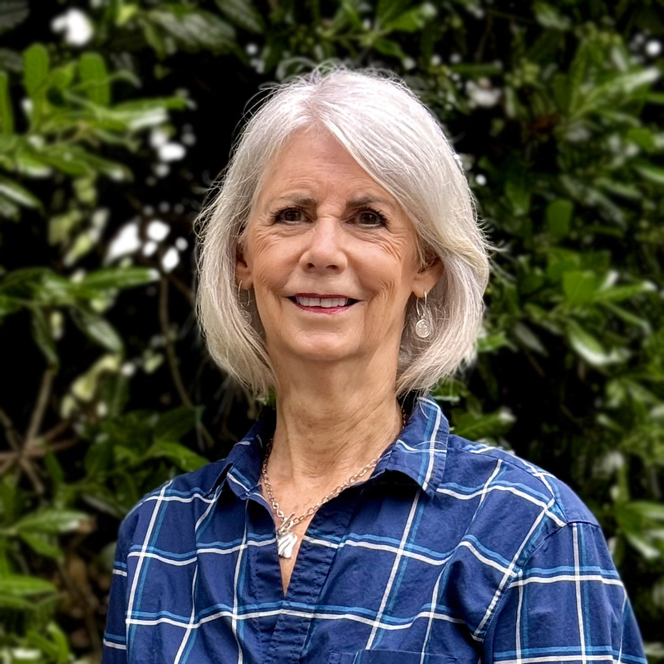 A smiling older woman with short gray hair wears a blue plaid shirt and a necklace, standing outdoors in front of green leafy plants.