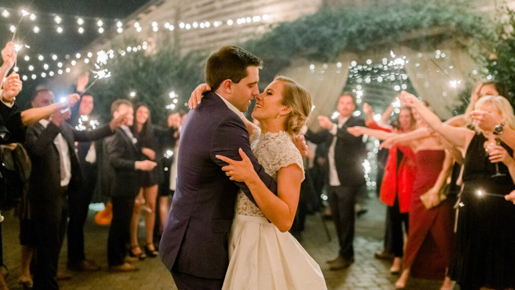 A bride and groom dance closely at an outdoor nighttime wedding reception, surrounded by guests holding sparklers and fairy lights, celebrating under glowing string lights.
