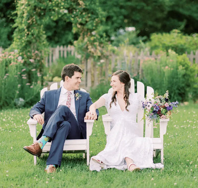 A bride and groom sit on white Adirondack chairs in a garden, smiling at each other. The groom wears a suit, and the bride wears a white dress, holding a bouquet of flowers. Greenery and flowers are in the background.