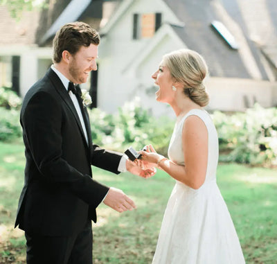 A groom in a black tuxedo and a bride in a white dress stand outside, smiling and laughing together as the bride receives a small gift box, with a house and greenery blurred in the background.