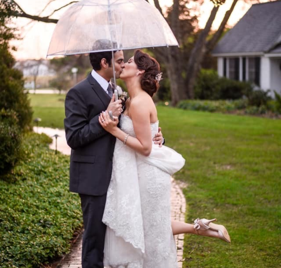 A bride and groom kiss under a clear umbrella in a garden, with the bride lifting one leg and holding onto the groom, both dressed in wedding attire. A house and trees are visible in the background.