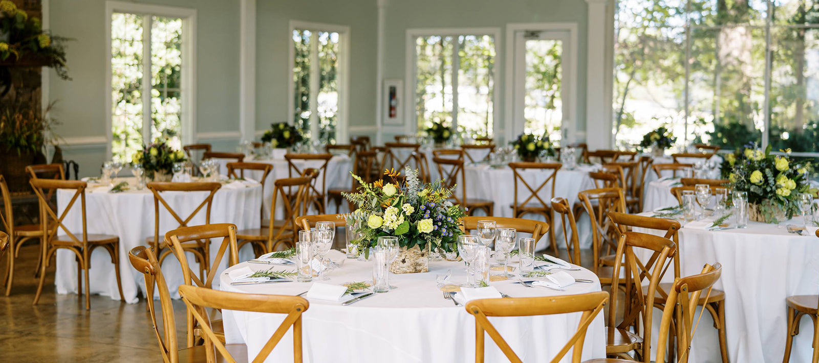 Round tables with white tablecloths and wooden chairs are set for an event in a bright room with large windows. Each table has a floral centerpiece and place settings with glasses and napkins. Sunlight fills the space.