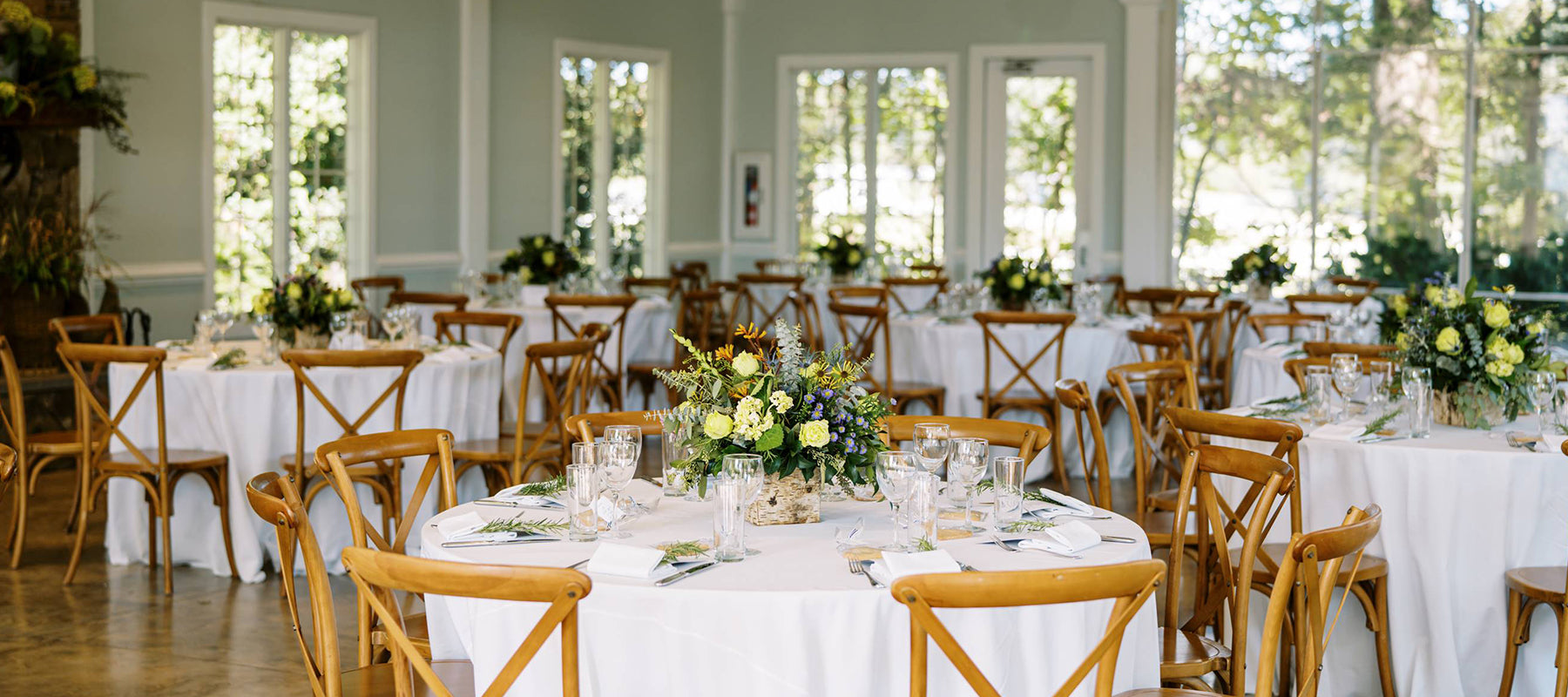 Round tables with white tablecloths and wooden chairs are set for an event in a bright room with large windows. Each table has a floral centerpiece and place settings with glasses and napkins. Sunlight fills the space.