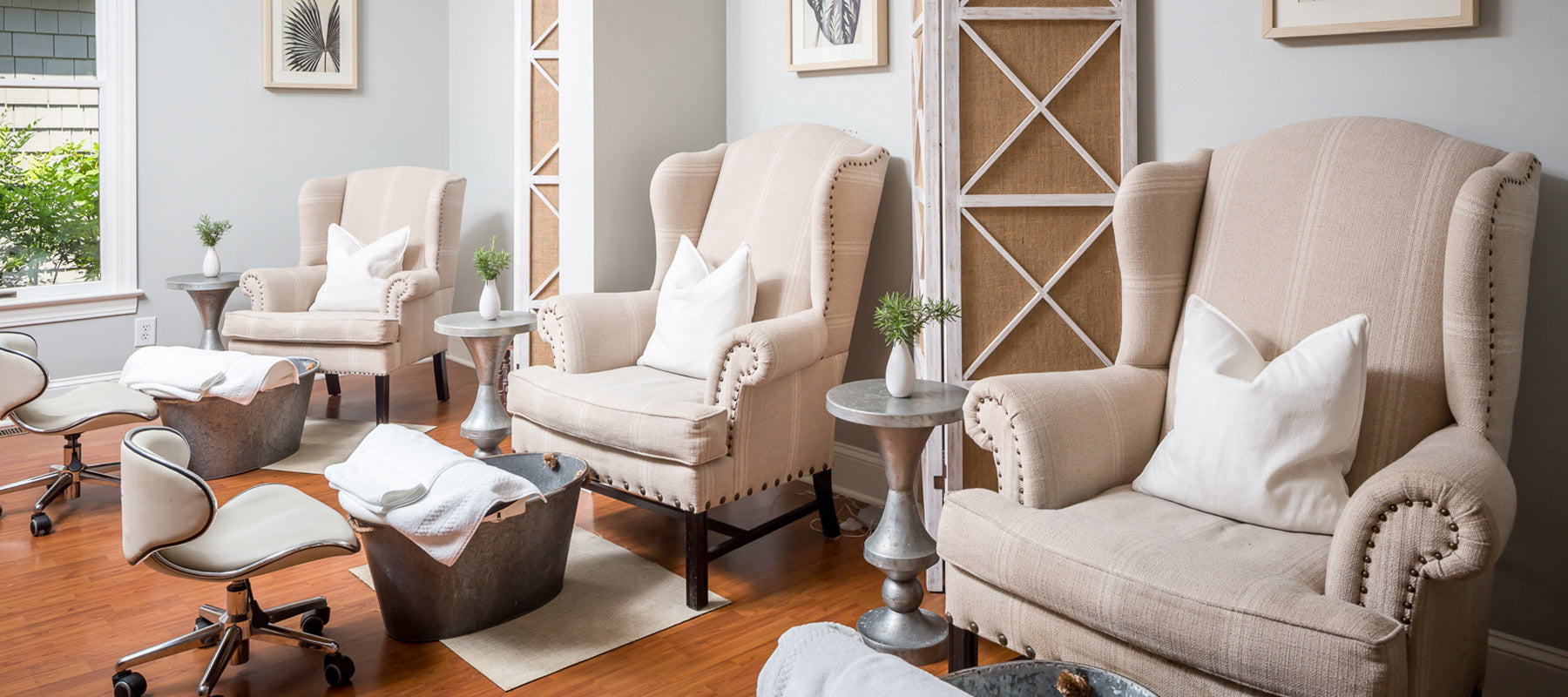 Three beige upholstered armchairs with white pillows are arranged in a bright, modern spa room. Each chair faces a foot spa tub and a rolling stool. The room features wood floors, small side tables, and framed art on the walls.