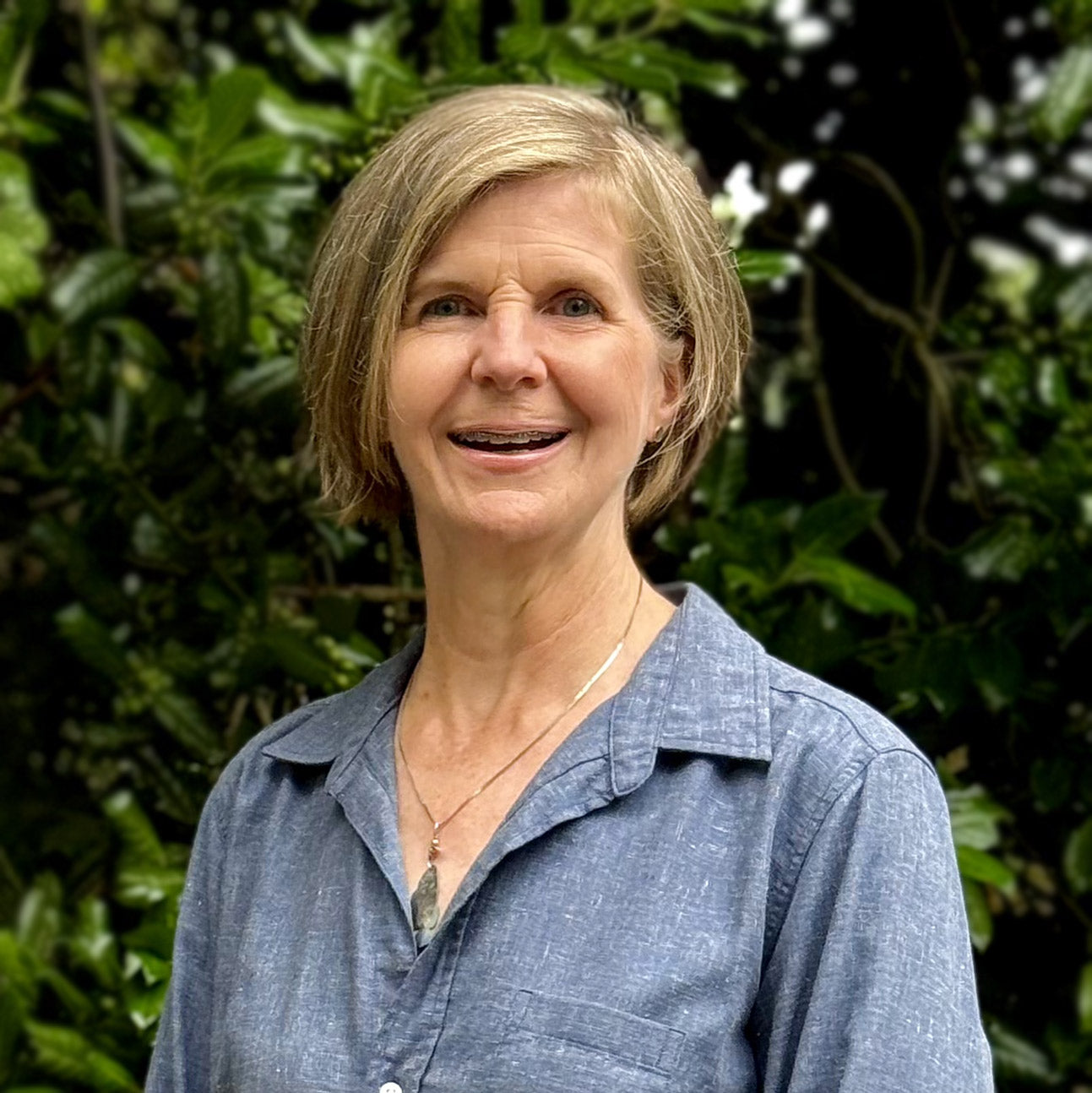 A woman with short blonde hair, wearing a blue collared shirt and a necklace, stands outdoors in front of leafy green foliage, smiling at the camera.