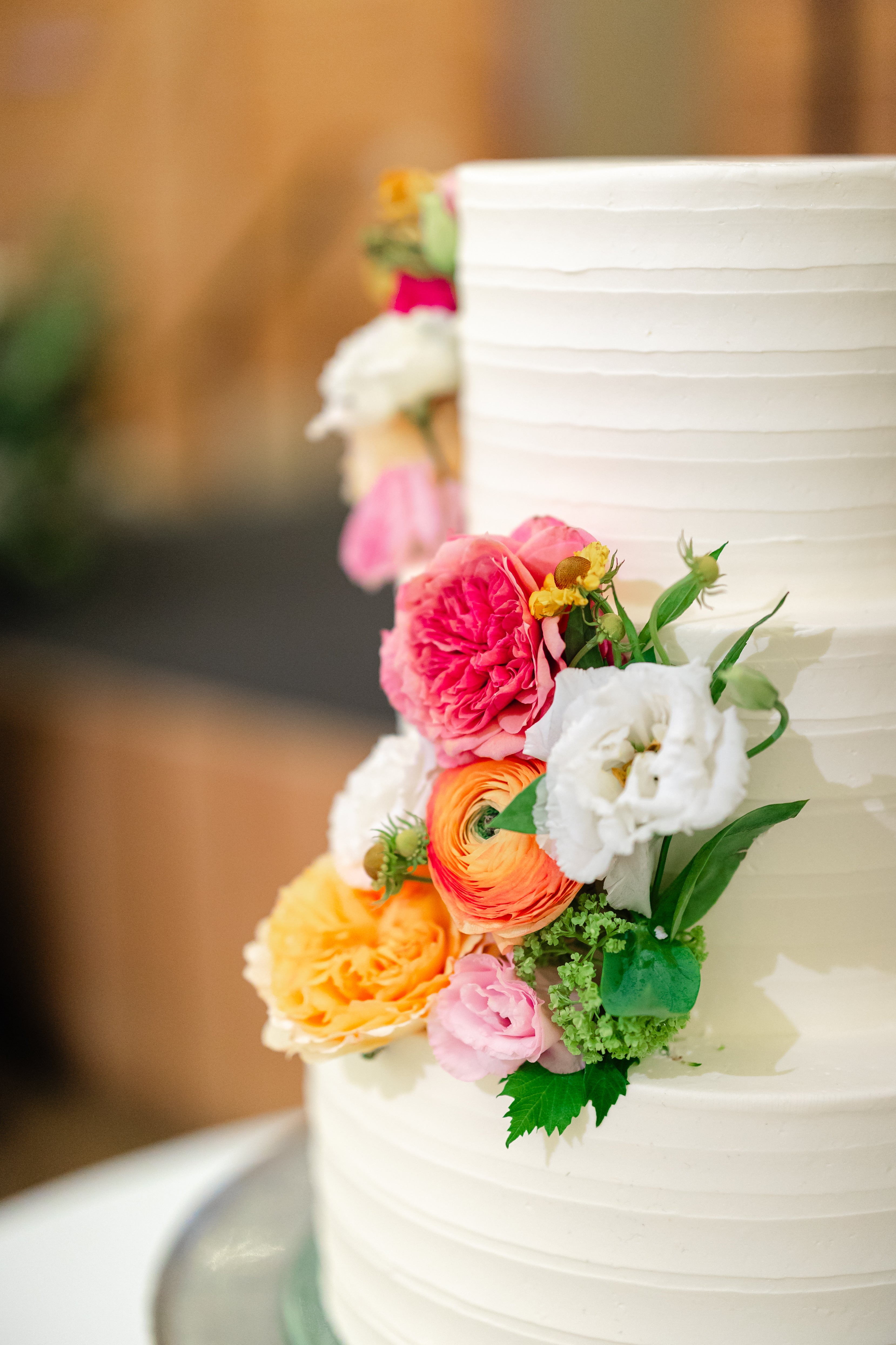 A white, tiered cake decorated with colorful fresh flowers, including pink, yellow, orange, and white blooms, sits on a table with a blurred background.