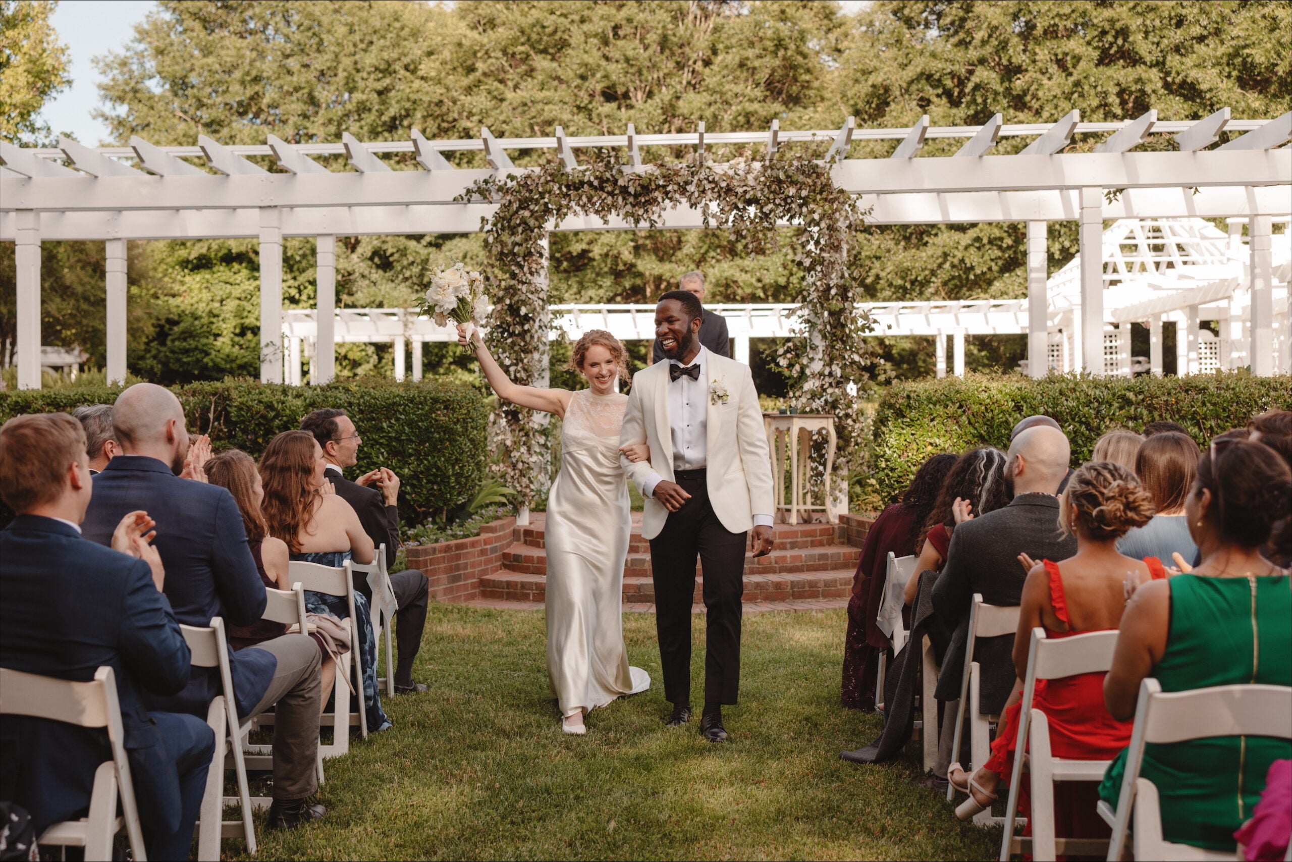 A joyful bride and groom walk down the aisle outdoors, smiling and holding hands, as guests seated on white chairs clap and celebrate their wedding under a white pergola decorated with greenery.