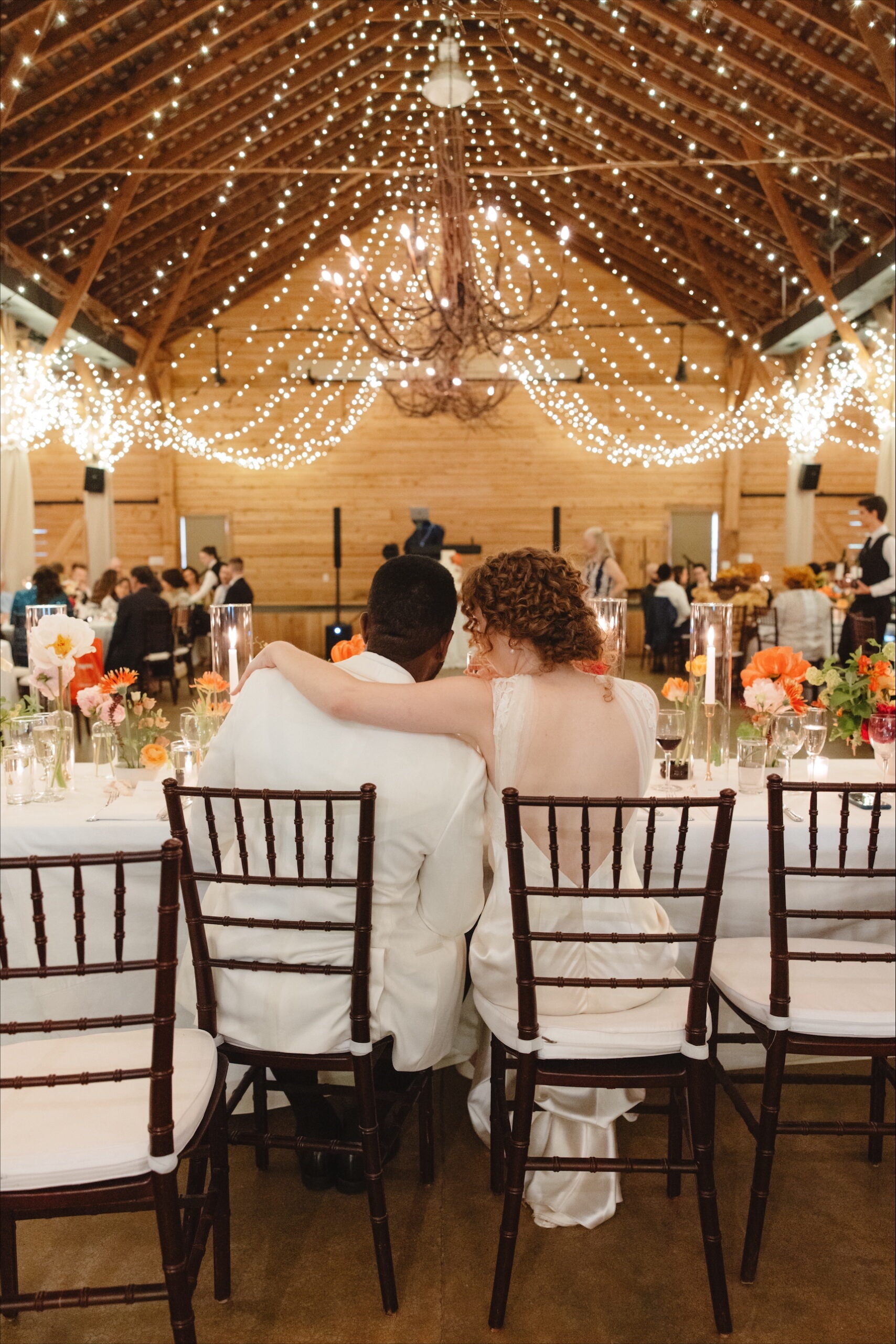A couple in white sits close together at a decorated table in a warmly lit barn wedding reception, surrounded by flowers and guests, beneath a ceiling draped with string lights.