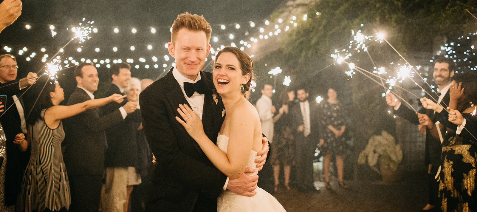 A bride and groom smile and embrace while wedding guests stand around them holding sparklers, with string lights glowing in the background at night.