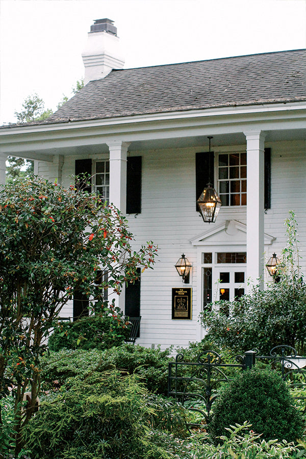 A white, two-story house with black shutters, tall white columns, and hanging lantern lights is surrounded by lush green bushes and trees. The entrance features a decorative pediment above the front door.