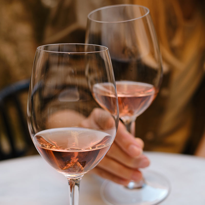 Two hands holding wine glasses filled with rosé, set against a softly blurred background, suggesting a relaxed, social atmosphere.
