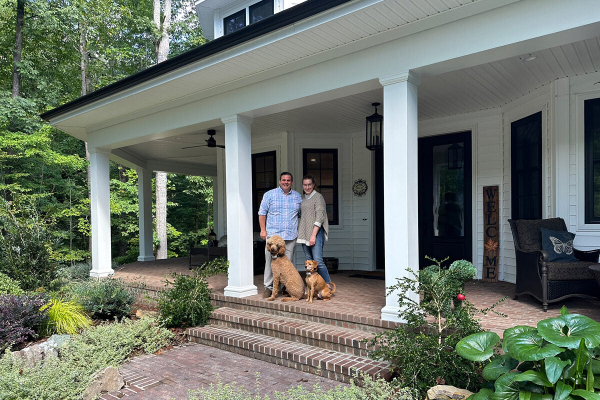 Two people stand on a brick porch in front of a white house, smiling at the camera. Two dogs sit beside them. The porch is surrounded by plants and trees, and there is a “Welcome” sign near the door.
