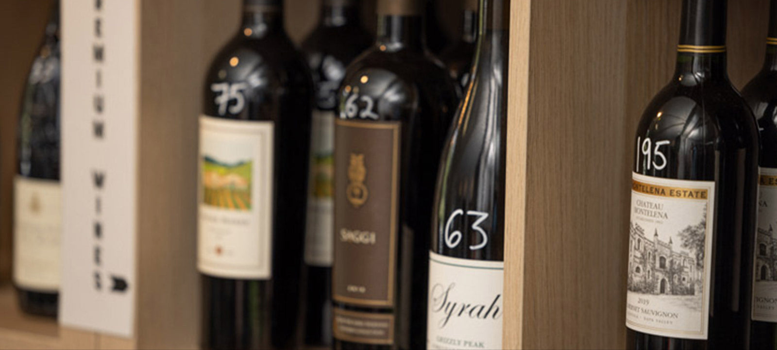 Several bottles of red wine are displayed upright on wooden shelves, with handwritten numbers like 75, 63, and 195 on the necks. The labels and shelving give a sense of an organized wine shop or cellar.
