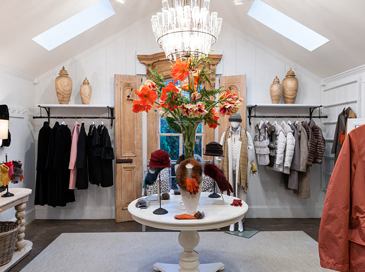 A stylish boutique displays coats and jackets on racks along white walls. A round table in the center holds hats, scarves, gloves, and a large bouquet of orange flowers beneath a chandelier.
