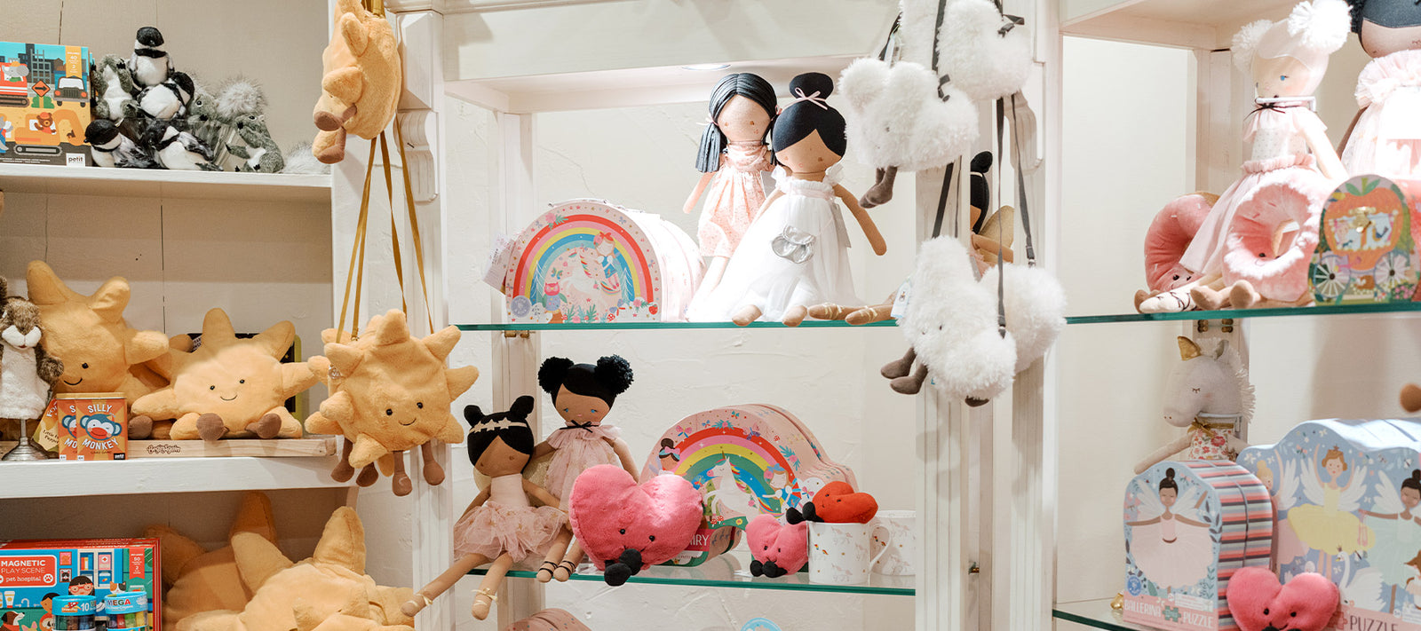 A display of plush toys, dolls, star-shaped pillows, heart plushies, and colorful rainbow decorations arranged neatly on glass shelves in a well-lit children’s store.