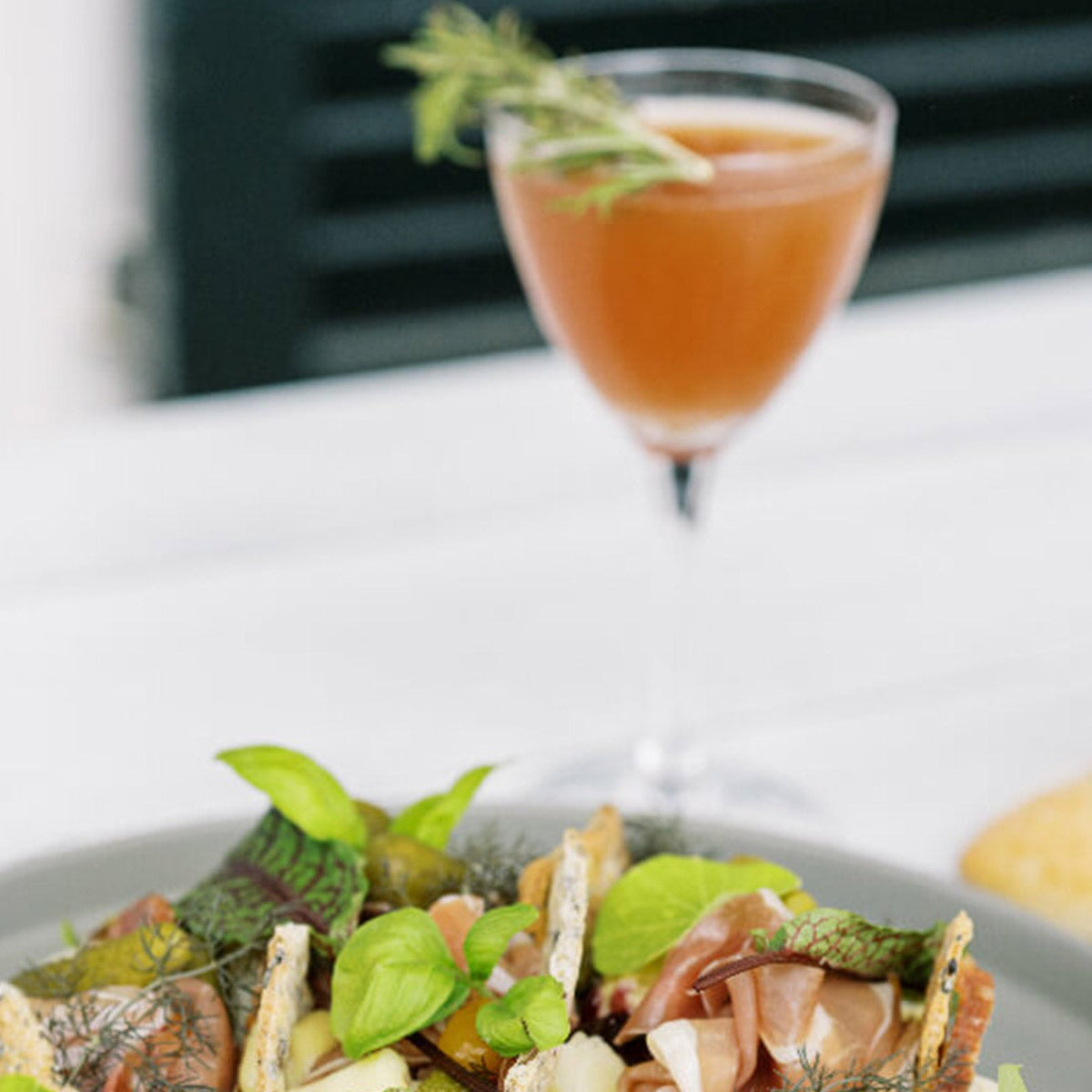 A close-up of a cocktail garnished with a rosemary sprig in a glass, set behind a plate of assorted fresh greens and appetizers, on a white table. The scene appears light and summery.