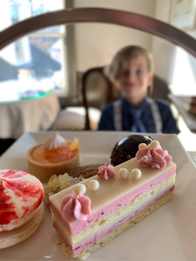 A plate of assorted pastries, including a layered pink and white cake with decorative icing, is in focus in the foreground. In the blurred background, a child in formal attire sits at a table.