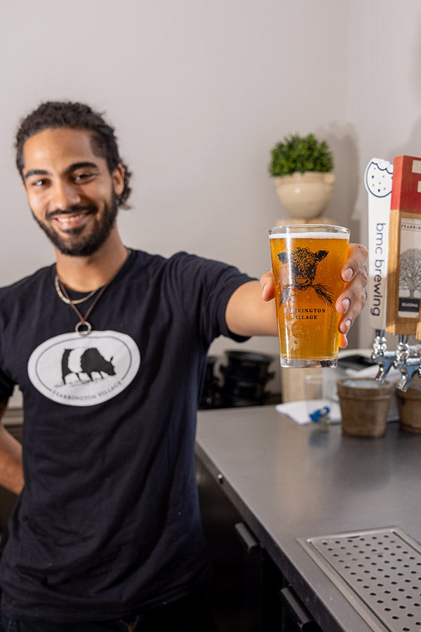 A smiling person with a beard and dark hair, wearing a black t-shirt, stands behind a bar and holds out a pint glass of amber beer toward the camera. Draft taps and a potted plant are visible in the background.