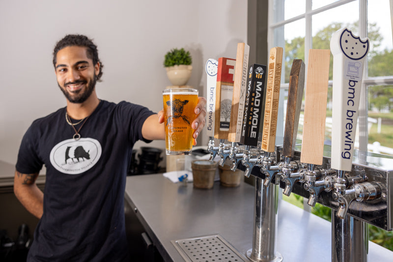 A smiling bartender with a beard and tied-back hair stands behind a bar, holding out a pint of beer. Several beer taps with different labels are visible, and sunlight streams in through a nearby window.