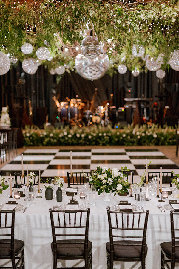 Elegant wedding reception setup with a white table, black chairs, green and white floral centerpieces, and a checkerboard dance floor. Hanging greenery and crystal chandeliers decorate the ceiling, with a stage and band in the background.