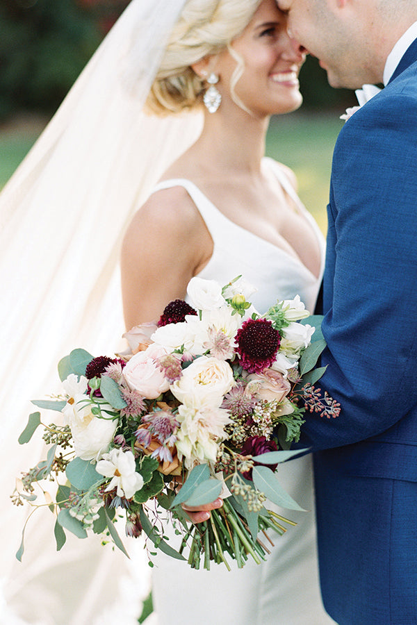 A bride in a white dress holds a large bouquet of pastel and dark flowers while standing close to a groom in a blue suit. They are smiling and touching foreheads, with the bride’s veil flowing behind her.